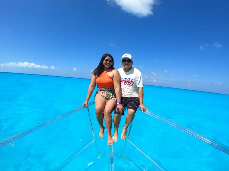 A happy couple sitting together on the front of a transparent boat, floating on the vibrant turquoise water of Cancun.