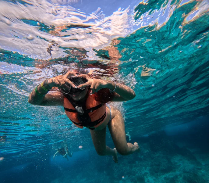 Mujer joven haciendo una señal de corazón con las manos mientras hace snorkel en las aguas turquesas de Cozumel durante el tour en barco transparente.