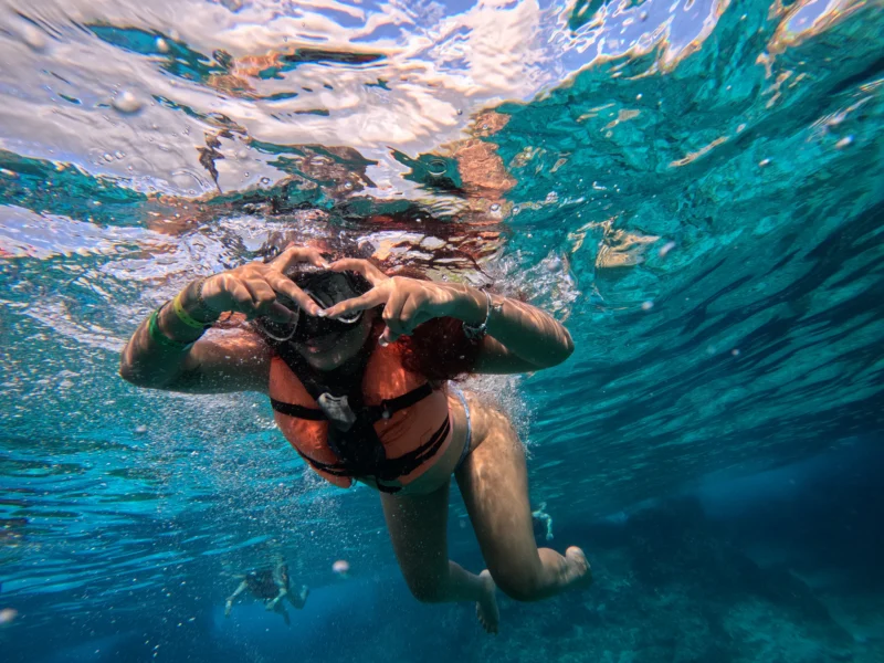 Mujer joven haciendo una señal de corazón con las manos mientras hace snorkel en las aguas turquesas de Cozumel durante el tour en barco transparente.