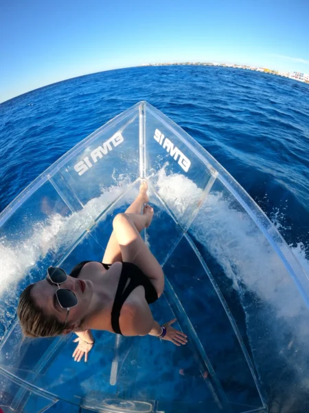 Wild Dolphins & The Transparent Boat: The Ultimate Cozumel Experience 🐬 3 Woman relaxing on a completely transparent boat while cruising over the deep blue sea of Cozumel, with the coastline visible on the horizon.