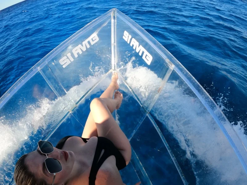 Woman relaxing on a completely transparent boat while cruising over the deep blue sea of Cozumel, with the coastline visible on the horizon.
