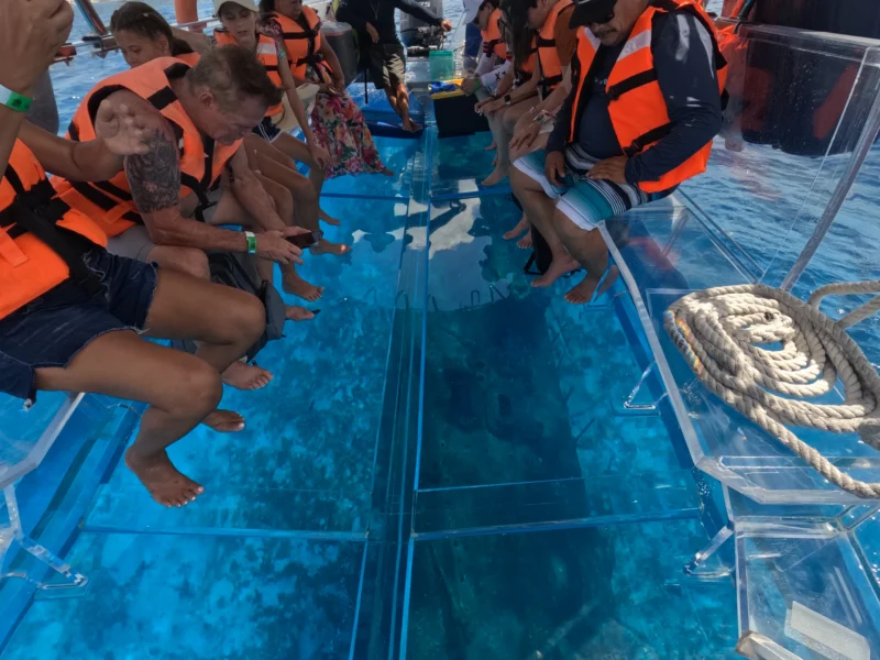 Grupo de turistas con chalecos salvavidas sentados en una lancha completamente transparente, observando el arrecife y el fondo marino a través del suelo de cristal.