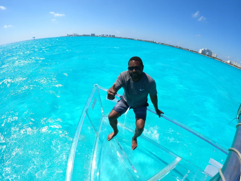 Hombre sonriendo y dando un pulgar hacia arriba mientras está sentado en un barco 100% transparente en las aguas de Cancún, con la zona hotelera al fondo.