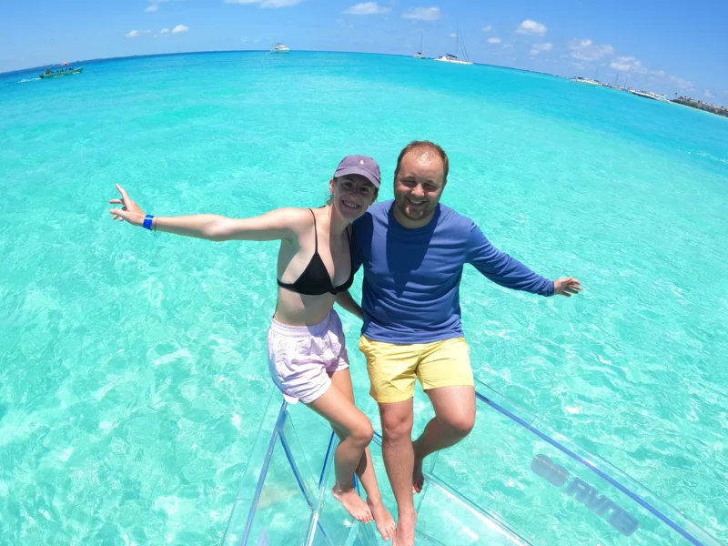 A happy couple laughing and enjoying the view from a 100% transparent boat on the crystal-clear turquoise waters of Isla Mujeres, Mexico.