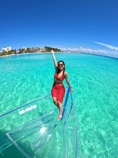 Happy woman posing on a 100% transparent boat, revealing the stunningly clear turquoise ocean and marine life below in Isla Mujeres, Mexico.