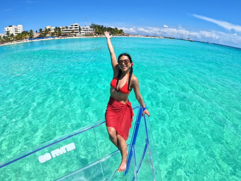 Happy woman posing on a 100% transparent boat, revealing the stunningly clear turquoise ocean and marine life below in Isla Mujeres, Mexico.