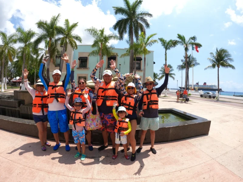 A large happy family, with children and grandparents, wearing life jackets and waving on the Cozumel waterfront before their clear boat tour.