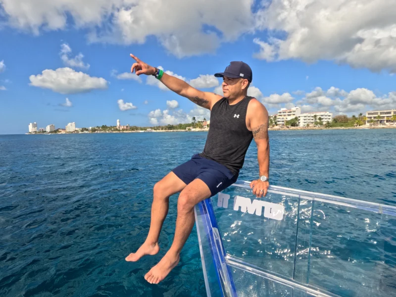 Hombre sentado en la proa de una lancha transparente durante un tour, apuntando hacia la costa en un día soleado.