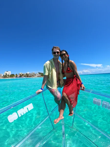 Pareja feliz disfrutando de un tour romántico en una lancha completamente transparente sobre el agua cristalina y turquesa de Isla Mujeres, México.