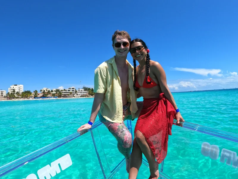 Pareja feliz disfrutando de un tour romántico en una lancha completamente transparente sobre el agua cristalina y turquesa de Isla Mujeres, México.