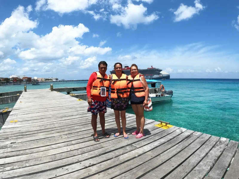 A happy family in life vests standing on a wooden pier, excited for their clear boat Cozumel tour with a cruise ship visible on the stunning turquoise sea. Near the Transparent Boat Cozumel Office