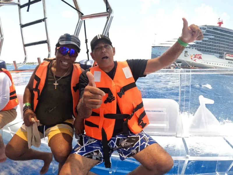 Dos turistas sonriendo y dando pulgares arriba mientras disfrutan de un tour en barco 100% transparente en Cozumel, con el mar Caribe y un crucero en el fondo.