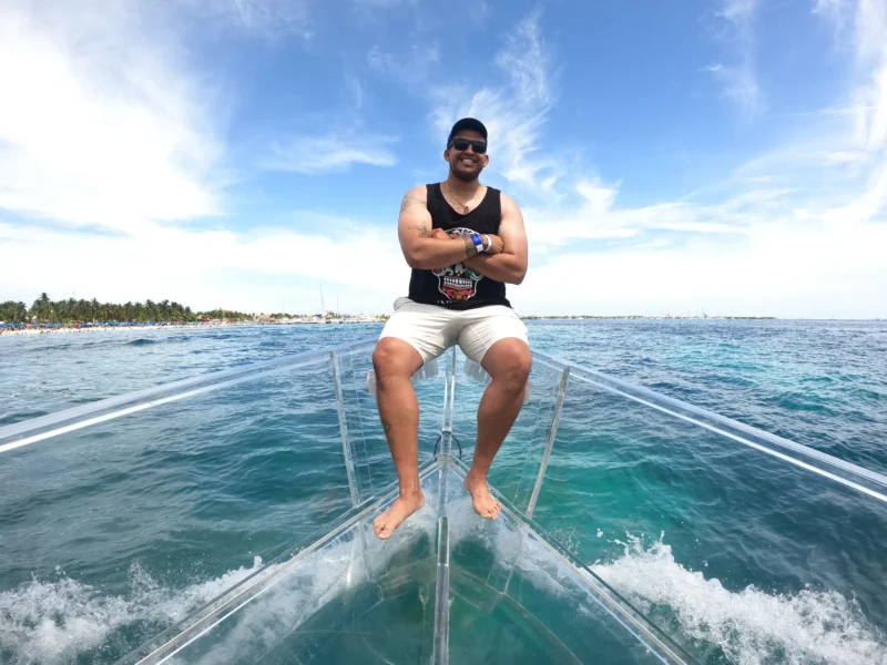 Hombre sonriendo y posando con confianza en la proa de un barco 100% transparente, navegando por las aguas turquesas de Isla Mujeres con Playa Norte de fondo.