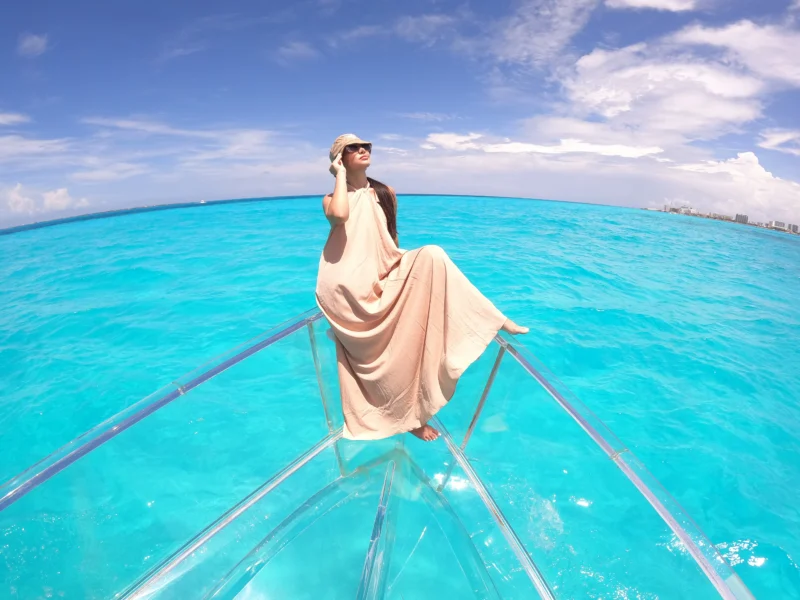 Woman in a flowing dress and sunglasses posing on the bow of a completely transparent boat, appearing to float over the brilliant turquoise water in Cancun.