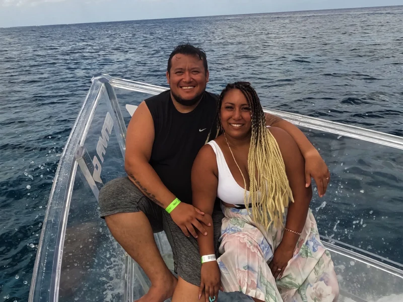 A happy couple smiling and embracing on the bow of a 100% transparent boat during a romantic clear boat Cozumel tour on the open sea.