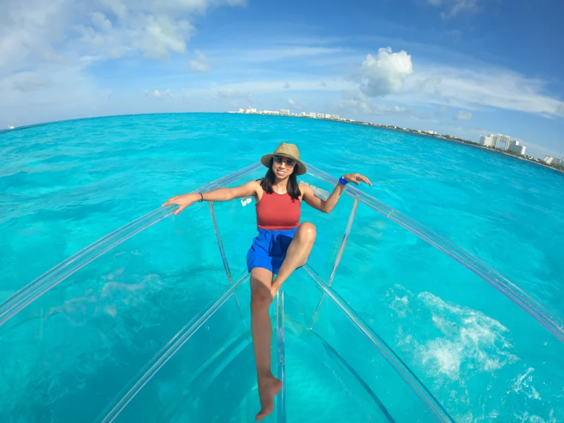 Happy woman with a sun hat smiling on the bow of a 100% transparent boat, enjoying the view of the Cancun skyline over the vibrant turquoise sea.