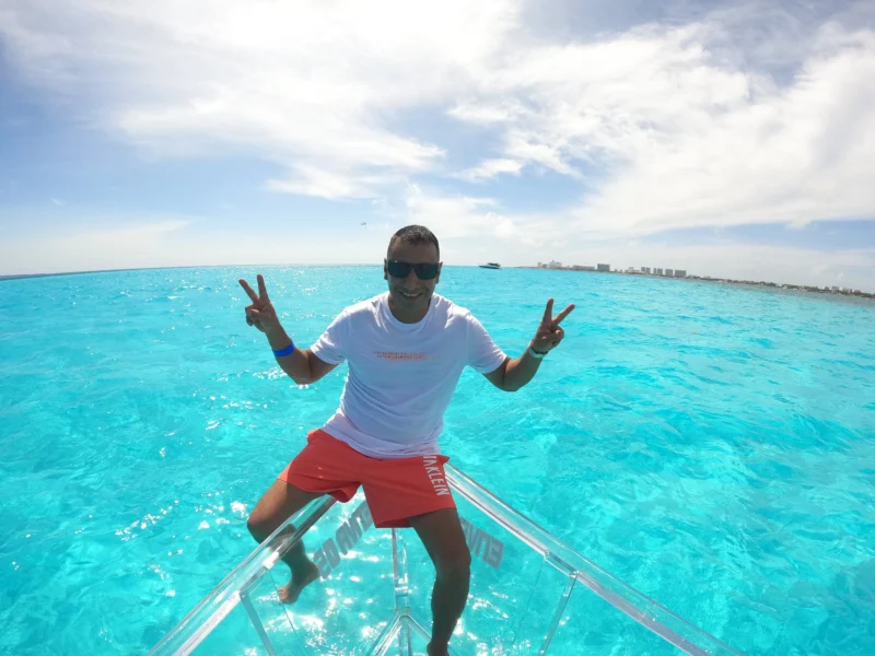 Hombre feliz con gafas de sol haciendo el símbolo de la paz en la proa de un bote 100% transparente sobre el increíble mar turquesa de Cancún.