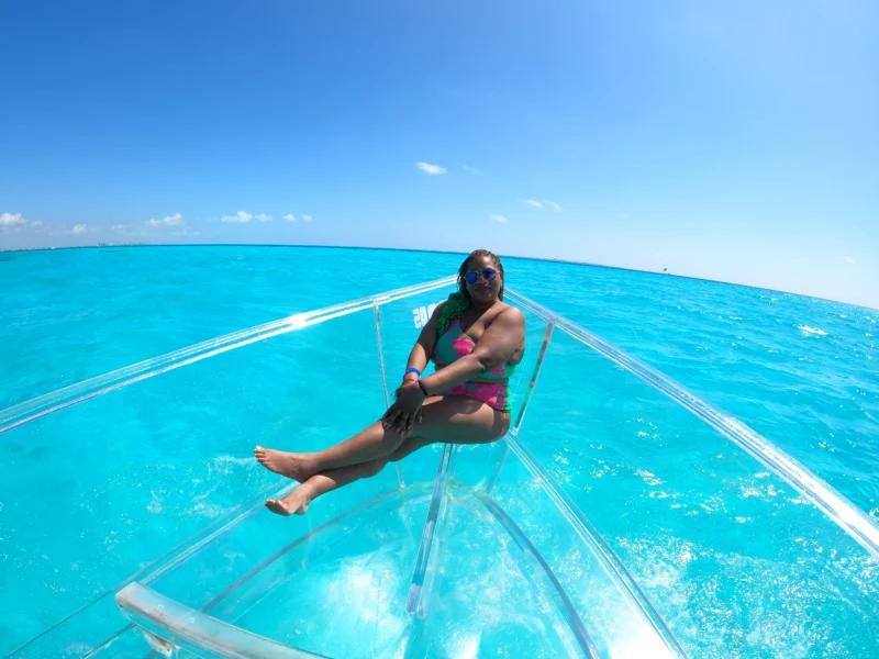 Woman relaxing on the bow of a clear boat in Cancun, surrounded by vibrant turquoise water under a clear blue sky.