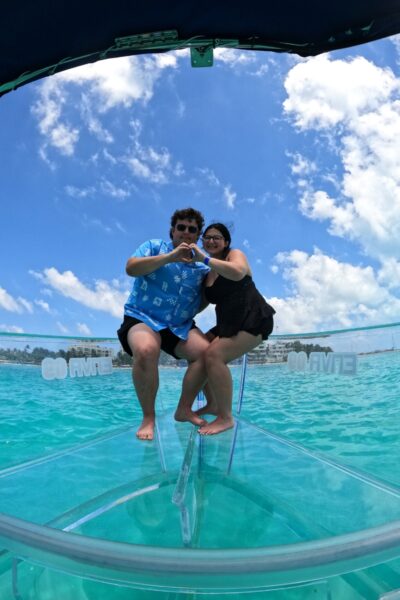 Una pareja haciendo la forma de un corazón con las manos en la proa de cristal de un barco transparente bajo un cielo azul en el Caribe.