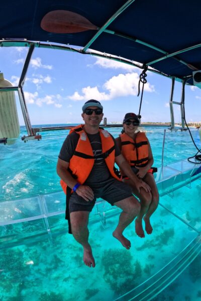 Pareja sonriente sentada en el borde de una lancha transparente sobre aguas turquesas cristalinas en Isla Mujeres.
