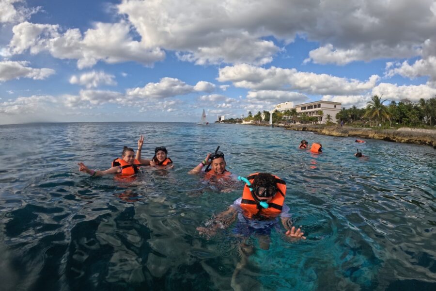 Group of friends and family wearing orange life jackets enjoying snorkeling in Cozumel ocean with the coast in the background
