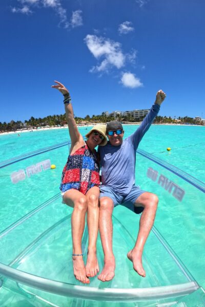 Dos turistas celebrando con los brazos en alto en la proa transparente de una lancha, mostrando el fondo marino de Isla Mujeres.