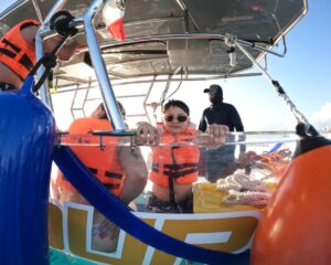 A young boy wearing sunglasses and a life jacket smiling at the camera while leaning on the transparent side of the boat, with a crew member in the background under the sunny Cozumel sky.