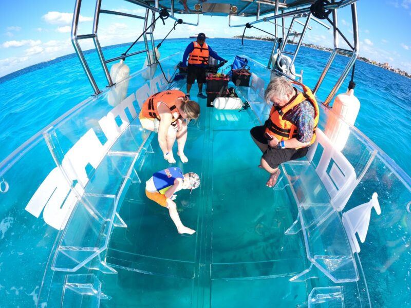 A young child in a blue life jacket looking through the 100% transparent glass floor of a boat at the turquoise water in Cozumel.