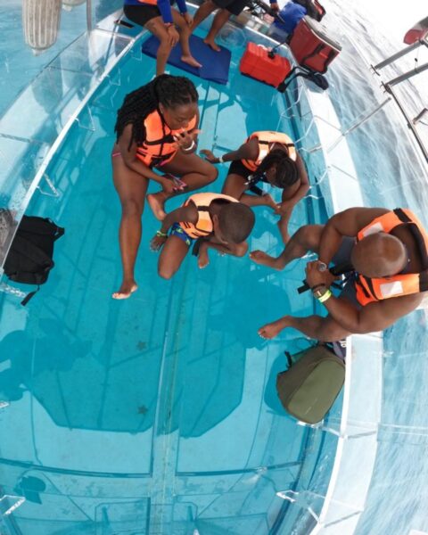 A family in orange life jackets sitting on the 100% transparent glass floor of a boat in Cozumel, looking at the clear blue water below.