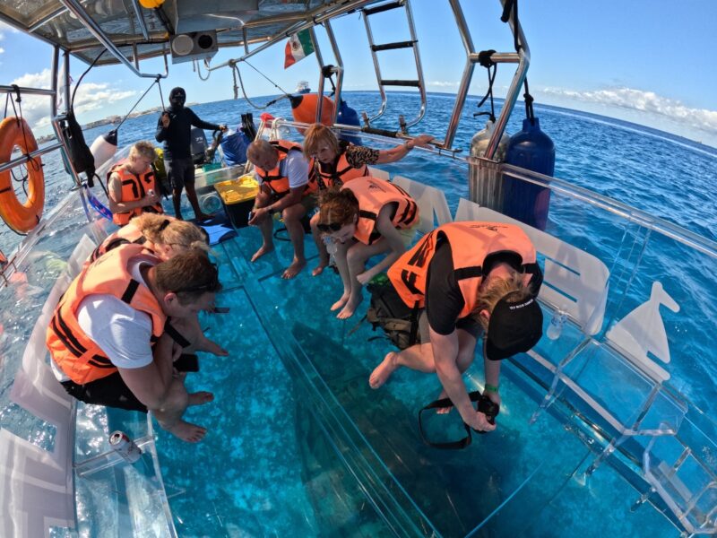 Tourists on a transparent boat in Cozumel photographing a sunken shipwreck visible through the glass floor.