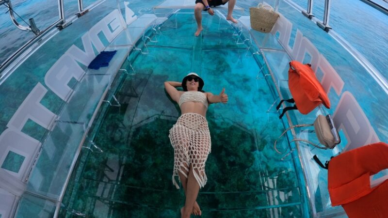 Luxury Clear Boat Cozumel tour showing a woman relaxing on a 100% transparent floor over a vibrant coral reef.