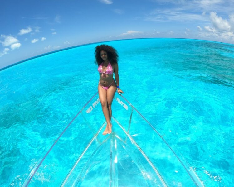 A beautiful woman in a pink bikini posing on a transparent boat in the shallow, turquoise waters of El Blanquizal, Cancun, optimized for Caribbean boat tours World Cup 2026.