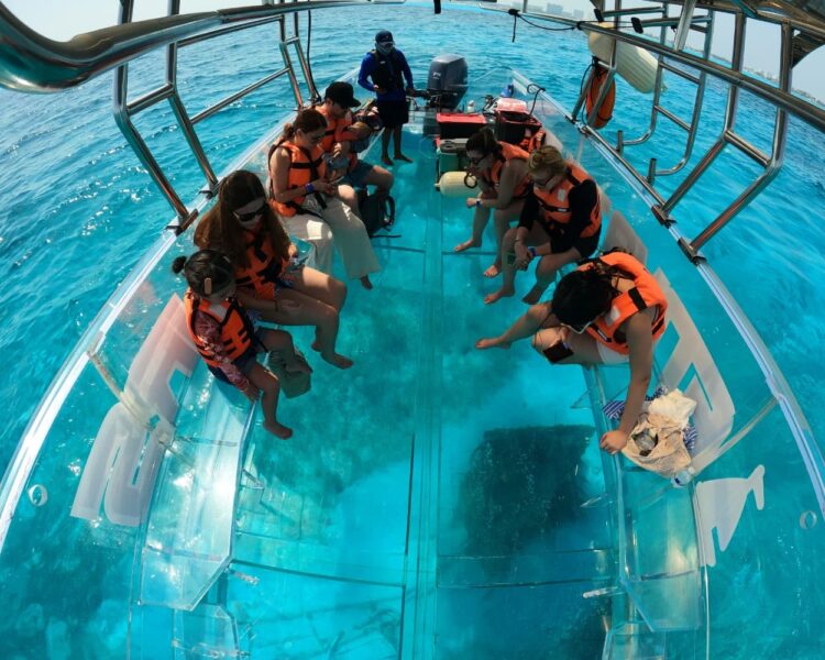 A group of tourists in life jackets sitting on a transparent boat and looking at a shipwreck on the ocean floor during a Caribbean Boat Tour for FIFA World