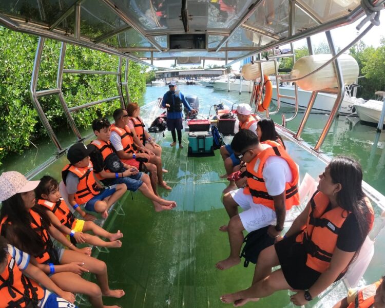 A group of ten happy tourists of diverse backgrounds wearing orange life jackets and sitting on a transparent boat posing for a photo on the clear water of Cancun during a Caribbean boat tour World Cup 2026 excursion in Mexico.