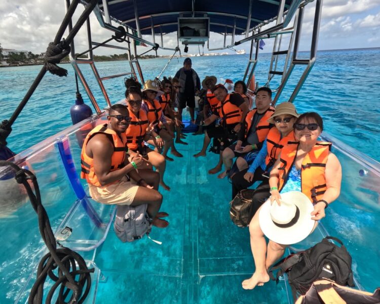 A group of ten happy tourists, of diverse backgrounds, wearing orange life jackets and sitting on a transparent boat, posing for a photo on the clear turquoise water of Isla Mujeres during a Caribbean boat tours World Cup 2026 in Mexico.