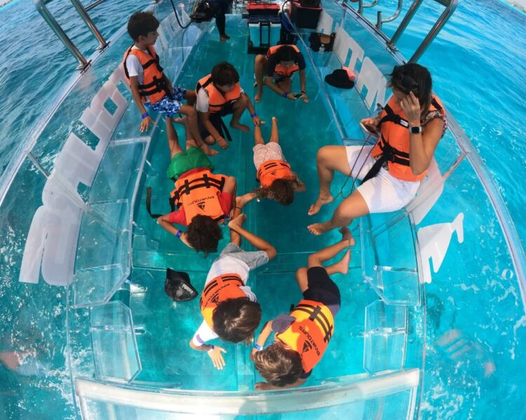 An overhead view looking down into a 100% transparent boat in Cancun, with a family of adults and children wearing orange life jackets lying on their stomachs and peering through the clear floor at the vibrant turquoise ocean water below.