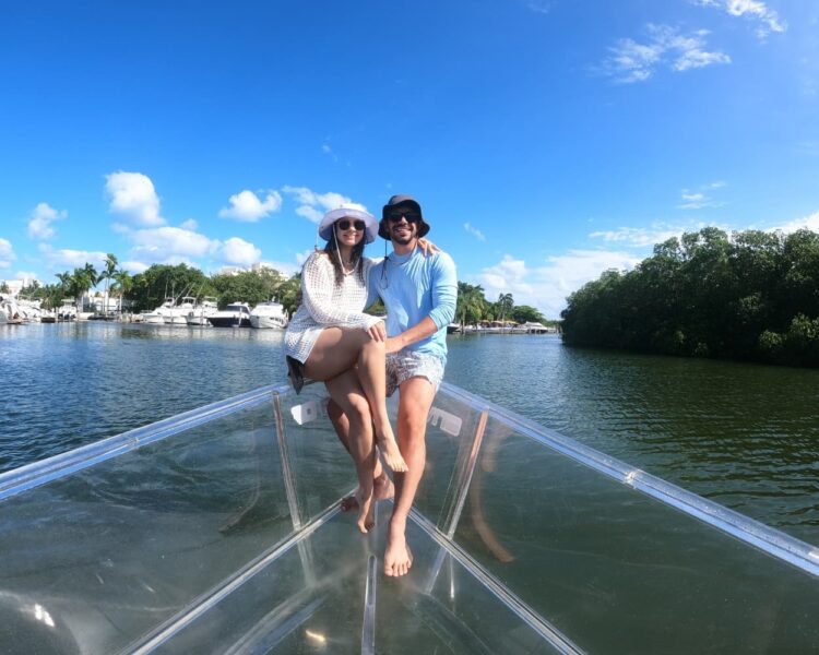 Clear Boat Cancun Tour: The Ultimate 100% Transparent Experience 🇺🇸 1 A happy couple sitting on the bow of a transparent boat during a Clear Boat Cancun Tour, with luxury yachts, palm trees, and the Nichupte lagoon mangroves in the background.