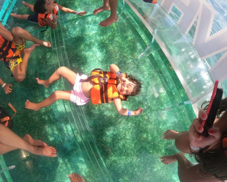A young girl smiling and making a peace sign while lying on the transparent floor of the Clear Boat Isla Mujeres Tour, looking at the green marine life below.