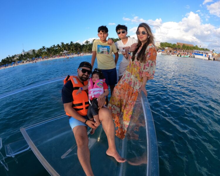 A family of five, including children and parents, posing on the bow of a transparent boat during a Clear Boat Isla Mujeres Tour, with the turquoise ocean and North Beach shoreline in the background.