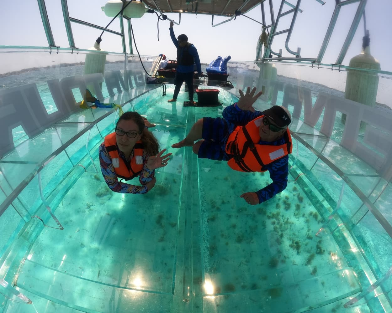 Two tourists wearing orange life jackets lying on the transparent floor of a clear boat isla mujeres tour, waving at the camera while looking at the sandy ocean floor in the crystal clear waters of Isla Mujeres.