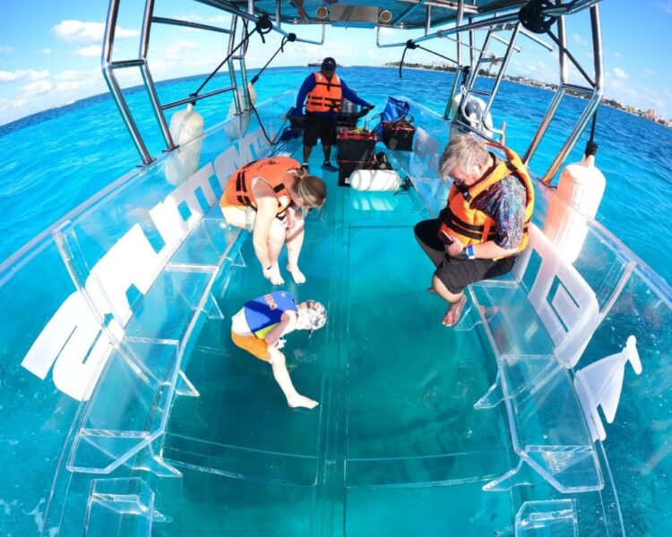 Tourists on a transparent boat looking down at a sunken shipwreck on the sandy ocean floor of Cozumel through the clear glass bottom.