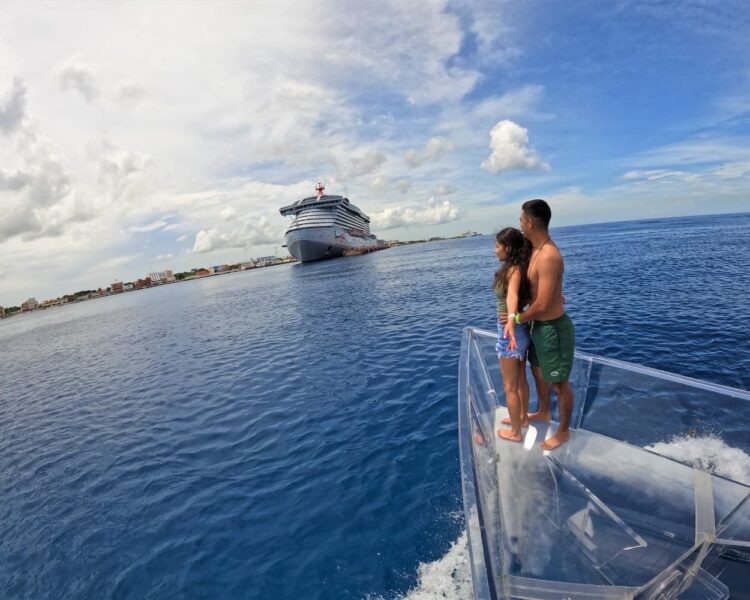 A young couple standing on the bow of a transparent boat, looking at a large cruise ship docked in the deep blue waters of Cozumel, Mexico.