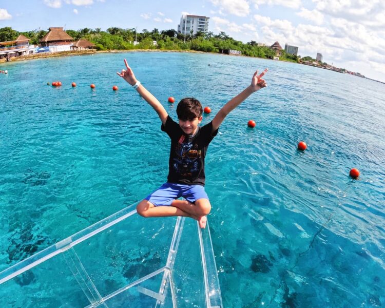 A happy boy sitting cross-legged with his arms up in a "rock on" gesture on the bow of a transparent boat, overlooking the vibrant turquoise ocean of Cozumel.