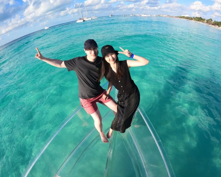 An overhead shot of a couple wearing hats, sitting on the front of a transparent boat with the turquoise ocean around them, making peace signs on their Clear Boat Isla Mujeres Tour.