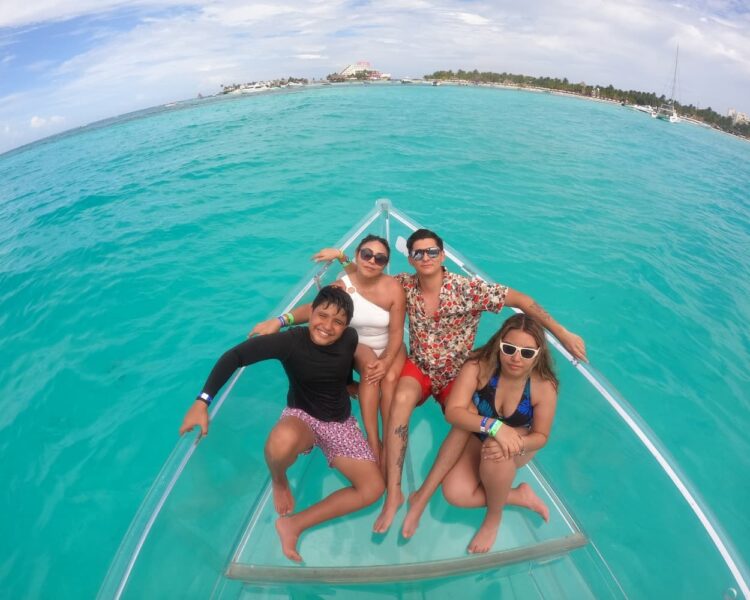 A group of four friends smiling and posing together on the bow of a transparent boat in the middle of the turquoise sea of Isla Mujeres.