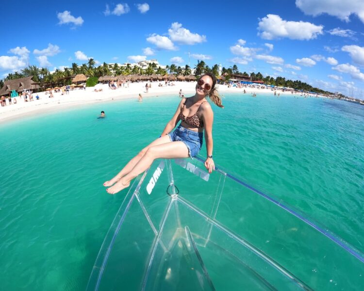 A woman in a leopard print bikini sitting on the bow of a transparent boat with the white sand and palm trees of North Beach Isla Mujeres in the background.