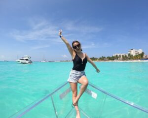 A happy woman in a black swimsuit and denim shorts posing excitedly with hands up on the bow of a transparent boat, with the turquoise waters and beachfront of Playa Norte, Isla Mujeres, in the background. courtesy clear boat tours