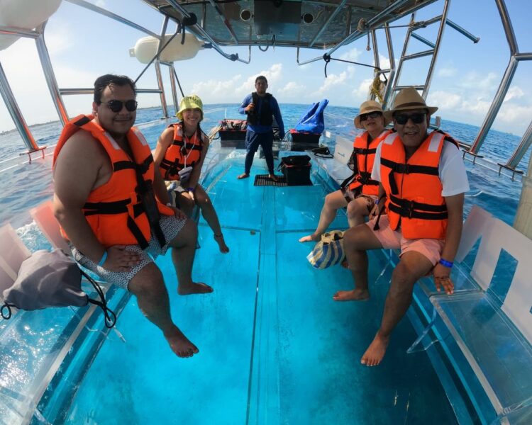 A group of four tourists wearing orange life jackets sitting comfortably inside a 100% transparent boat, with the captain in the background, navigating the turquoise waters of Isla Mujeres.