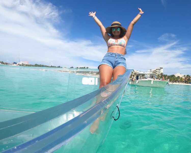 A woman with a sun hat and sunglasses raising her arms in joy while sitting on the edge of a transparent boat, with the turquoise water of Isla Mujeres and Playa Norte in the background.